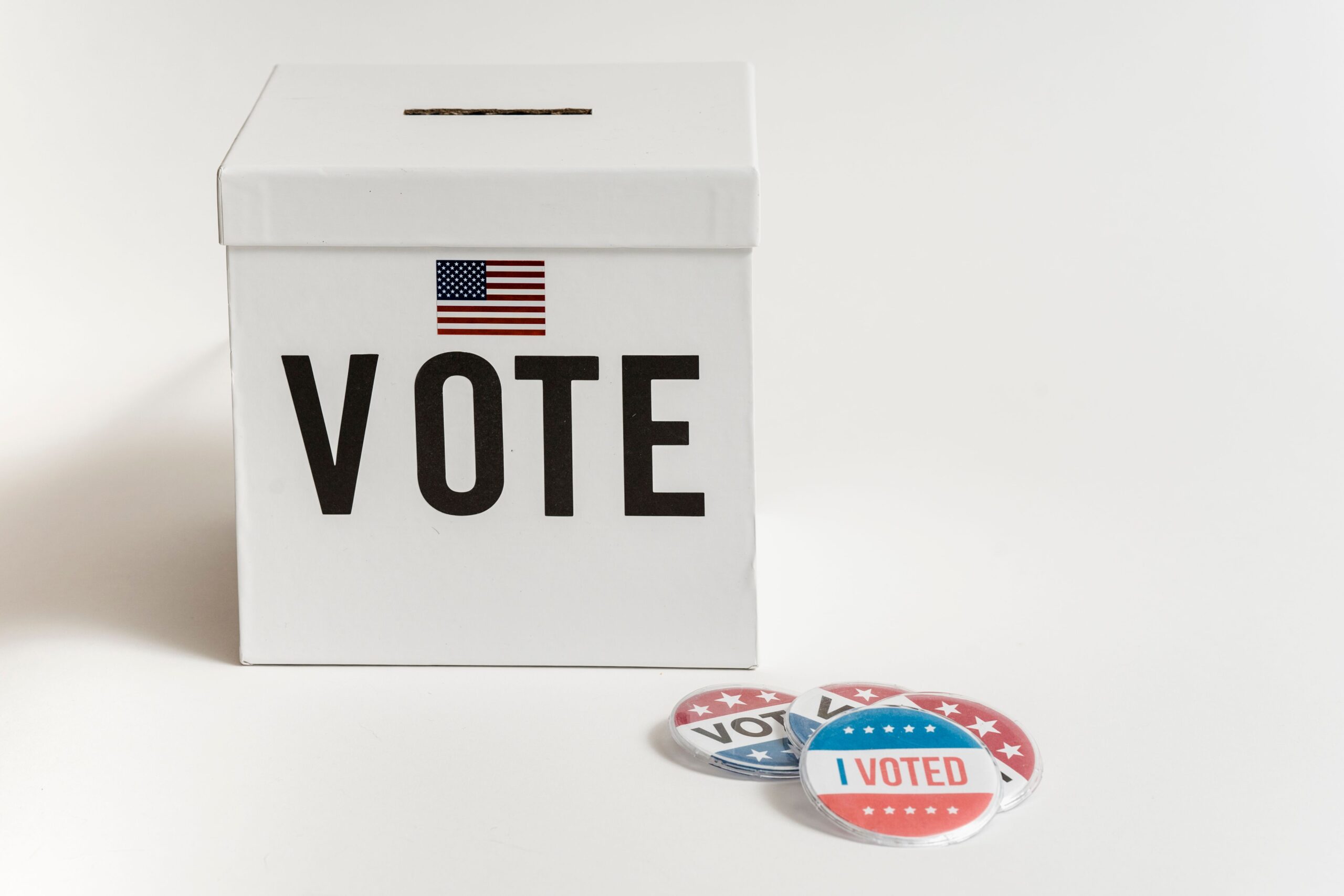 White ballot box labeled "VOTE" with small U.S. flag and "I Voted" buttons.
